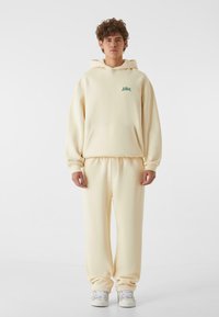 Cremefarvet hoodie og matchende sweatpants med en frontlomme, ribbede manchetter og et diskret grønt logo på brystet.
