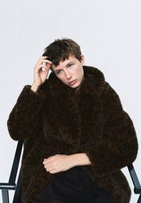 Cappotto in pelliccia sintetica marrone con stampa leopardata, dotato di colletto alto e chiusura a bottoni. Modella seduta su una sedia nera, con uno sfondo semplice.