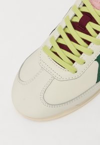 Baskets en cuir crème avec des accents verts, lacets texturés vert lime et une étiquette en violet sur la languette. Présente des coutures détaillées et une semelle en caoutchouc.