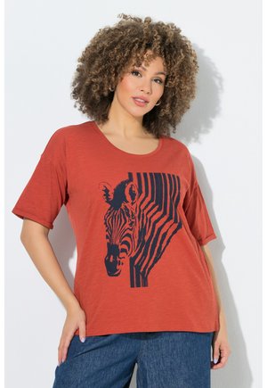 Frau mit lockigem Haar, die ein rostfarbenes T-Shirt mit einem schwarzen Zebra-Motiv trägt, kombiniert mit blauen Jeans, stehend vor einer weißen Wand.