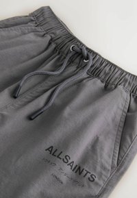 Pantalon en tissu gris avec taille élastique et cordon de serrage, arborant le logo "ALLSAINTS LONDON" et un texte japonais sur la cuisse.