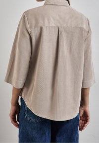 Camicia beige, leggera, con vestibilità comoda, maniche a 3/4 e spalla posteriore. Presenta un orlo curvo e una texture delicata.