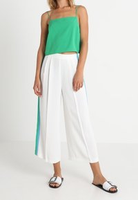 Débardeur vert court à fines bretelles, associé à un pantalon blanc à jambes larges avec deux rayures verticales bleu sarcelle. Sandales décontractées à enfiler.