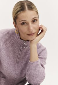 Ljust lila stickad tröja med hög krage, som har en silverfärgad dragkedja och ribbade ärmslut. Guldörhängen synliga. Slät textur.