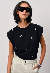 Zwarte top met korte mouwen en decoratieve bloemversieringen, gecombineerd met lichtgekleurde, hoge taillebroeken. Model draagt oversized zonnebril.