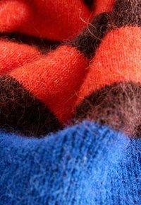 Fokussēts skats uz mīkstu, pūkainu adījumu ar sarkaniem, brūniem un ziliem horizontāliem svītrainiem, kas parāda tekstūru un šķiedru detaļas.