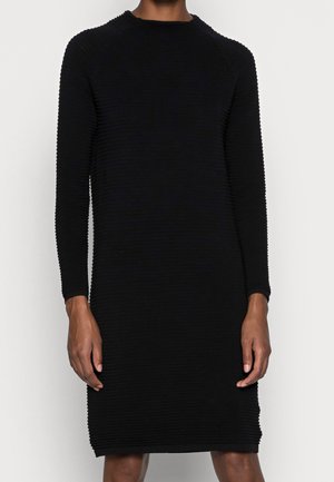 Person wearing a long-sleeve, black ribbed knit dress, knee-length, standing against a plain light background.