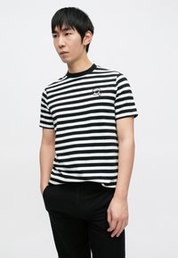 Czarna i biała koszulka w paski z okrągłym dekoltem, z małą grafiką na piersi. Tkanina wydaje się miękka i lekko teksturowana.