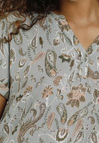 Blouse gris clair avec motifs paisley et floraux dans des tons verts doux, pêche et bruns ; texture douce ; col en V, manches courtes.