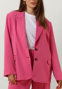 Overzised roze blazer met twee knopen, notch-lapels en zijzakken, gedragen over een wit T-shirt. Glad stof met een getailleerde pasvorm.