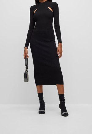 Frau trägt ein figurbetontes schwarzes Midikleid mit langen Ärmeln und vorderen Ausschnitten, hält eine kleine schwarze Handtasche in einer Hand.