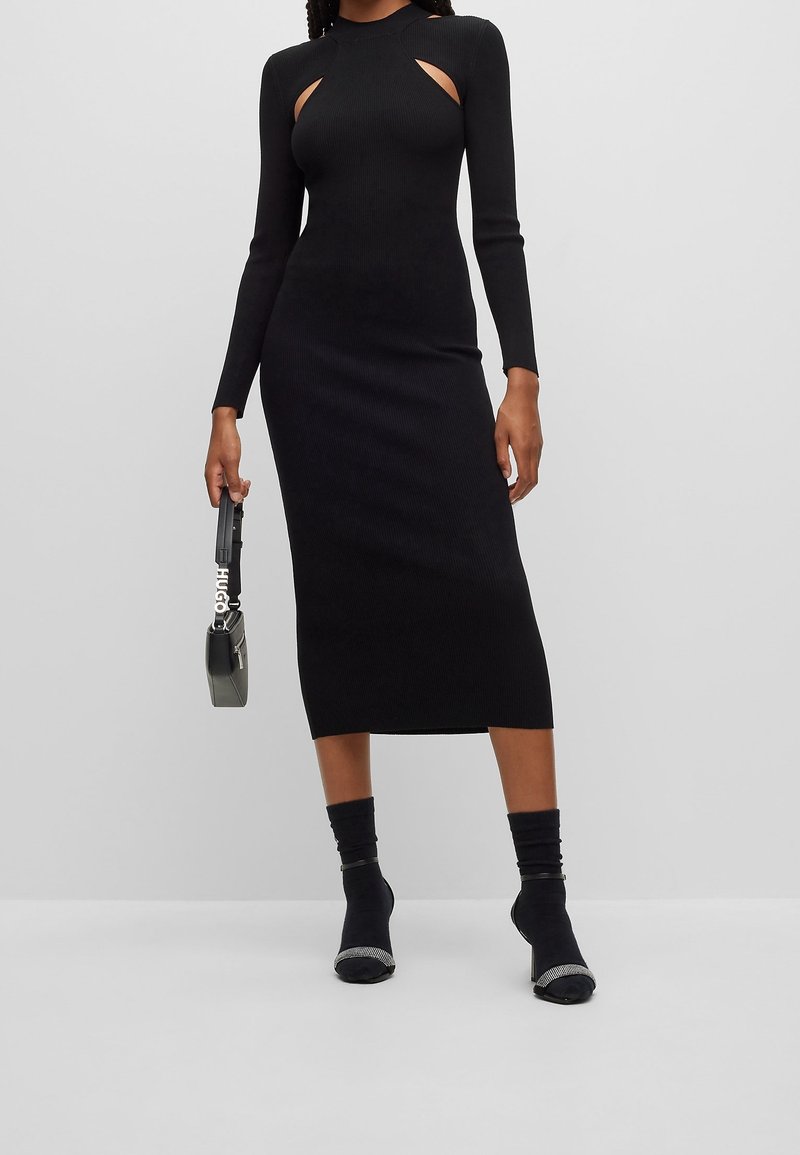 Femme portant une robe midi noire moulante avec des manches longues et des découpes à l'avant, tenant un petit sac à main noir dans une main.