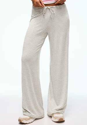Personne portant un pantalon large à rayures grises et blanches, taille avec cordon, et des baskets beige et blanc, les mains ajustant la taille.