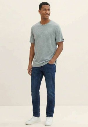Homme portant un t-shirt gris clair à manches courtes, un jean bleu foncé et des baskets blanches, debout avec une main dans la poche, souriant, sur un fond uni.