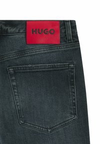 Jeans in denim scuro con un'etichetta in pelle rossa su cui è impresso "HUGO" in lettere nere in rilievo. Include una tasca posteriore e cuciture dettagliate.