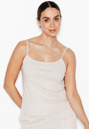 Donna con capelli scuri raccolti in uno chignon, indossa piccoli orecchini a cerchio e una canottiera beige chiaro, in piedi davanti a uno sfondo bianco semplice.