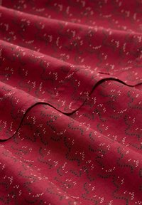Roter Stoff mit einer weichen Textur, der ein wirbelndes Muster aus schwarzen und weißen Punkten zeigt, mit sichtbaren gefalteten Kanten und Nähten.