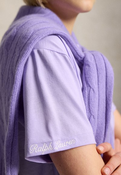Άτομο που φοράει μια λιλά μπλούζα Ralph Lauren με κεντημένο λογότυπο στο μανίκι και ένα ταιριαστό πλεκτό πουλόβερ περασμένο πάνω από τους ώμους.