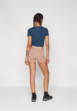 Femme aux cheveux tressés portant un t-shirt Nike bleu marine, un short Nike beige, des chaussettes noires et des baskets noires, debout de dos sur un fond uni.