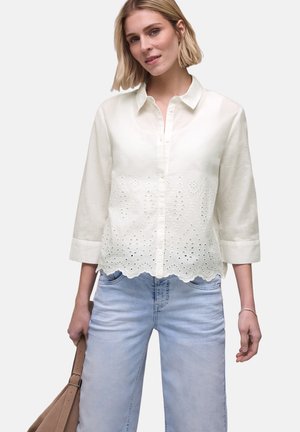 Kvinde iført en hvid bluse med lange ærmer med eyeletbroderi og bølget kant, lyseblå jeans, holder en lysebrun håndtaske.