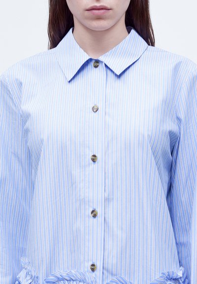 Femme portant une chemise boutonnée à rayures verticales bleu clair et blanc, avec un col pointu et un détail volanté en bas.