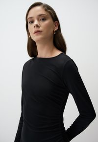 Haut noir à manches longues en tissu lisse et extensible, avec un col rond et des détails froncés sur les côtés.