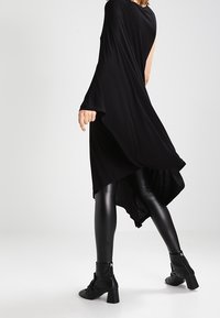 Svart asymmetrisk klänning med draperad design, som matchas med glänsande svarta leggings och ankelstövlar. Slät yta, flödande tyg och minimala detaljer.