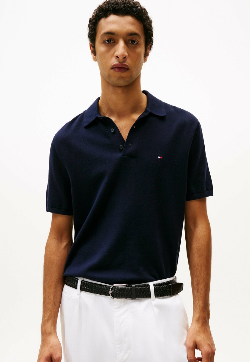 Homme portant un polo bleu marine et un pantalon blanc avec une ceinture noire tressée, debout devant un fond clair uni.