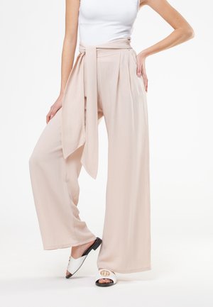 Donna che indossa pantaloni beige a gamba larga con cintura abbinata annodata in vita e sandali bianchi aperti con dettagli dorati.