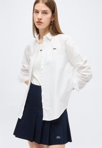 Νεαρή γυναίκα που φοράει λευκό πουκάμισο με κουμπιά πάνω από λευκό τοπ και μπλε σκούρα πλισέ φούστα με πράσινο λογότυπο κροκοδείλου.