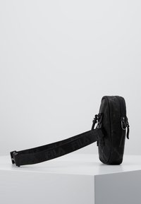 Czarna torba z teksturowanym wzorem, wyposażona w zamek błyskawiczny i regulowany pasek. Wykonana z trwałego materiału, kompaktowa i prostokątna w kształcie.