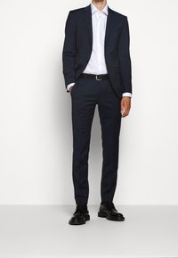 Tmavě modrý oblek s úzkým střihem, texturovaná látka, bílá košile pod ním, černý pásek a černé šněrovací boty.