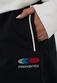 Main dans une poche de pantalon noir avec bordure blanche, texte brodé "PROHIBÉ" et formes ovales rouges, bleues et grises sur le tissu.