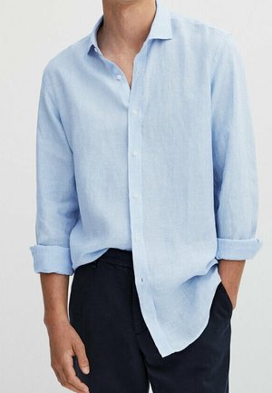 Homme portant une chemise bleu clair à manches longues boutonnée, manches retroussées, et un pantalon foncé, debout avec une main dans la poche.