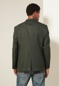 Blazer verde scuro in tessuto strutturato, con spacco singolo sul retro e polsini con bottoni, abbinato a jeans blu.