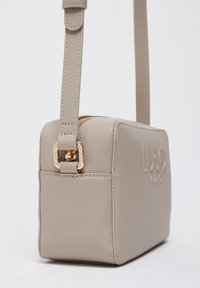 Beige synthetische handtas met ritssluiting, logodetail en gouden metalen accenten. Voorzien van een afneembare schouderriem. Rechthoekige vorm.