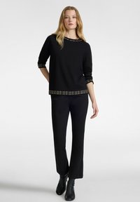 Top negro de manga larga con acentos en relieve de tachuelas doradas en el escote y el dobladillo, combinado con pantalones negros de campana y botines negros.