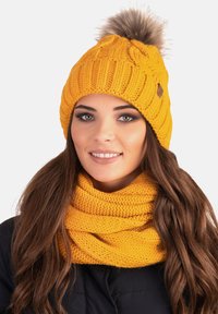 Mustard yellow knitted beanie with a textured pattern and a faux fur pom-pom on top, paired with a matching knitted scarf.