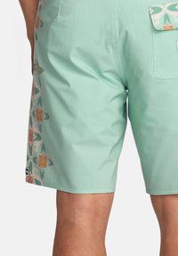 Pantaloni shorts verde menta realizzati in materiale leggero, con un motivo floreale colorato lungo il lato e una tasca posteriore con una pattina decorativa.