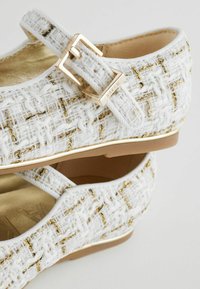 Weiße Stoffschuhe mit strukturiertem Finish, ausgestattet mit goldenen Akzenten und einem Schnallenriemen. Das Design beinhaltet eine runde Spitze und eine flache Sohle.
