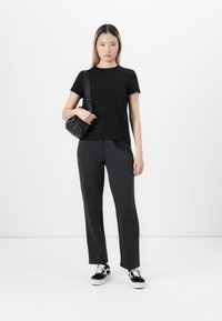 Jeune femme debout en t-shirt noir et pantalon, portant des baskets noir et blanc, tenant un sac à bandoulière noir contre un fond blanc.
