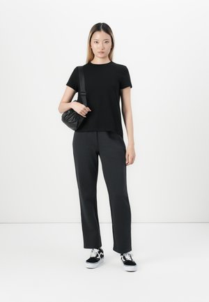 Jeune femme debout en t-shirt noir et pantalon, portant des baskets noir et blanc, tenant un sac à bandoulière noir contre un fond blanc.