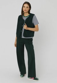 Jeune femme portant un gilet en polaire noir sur un t-shirt rayé, un pantalon noir à jambes larges et des baskets vertes et roses, se tenant devant un fond uni.