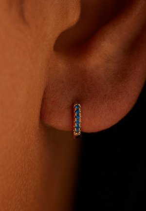 Ear with a small gold hoop earring set with five blue gemstones, against a dark background.