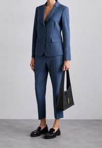 Completo su misura blu con giacca aderente e pantaloni affusolati; abbinato a mocassini neri e a una borsa nera strutturata. Finitura in tessuto liscia.