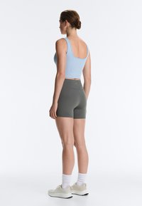Woman standing facing left, wearing a light blue sleeveless crop top, gray high-waisted shorts, white socks, and beige sneakers.