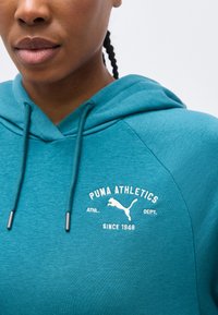 Persoană care poartă un hanorac turcoaz Puma Athletics cu logo și text alb, cu păr împletit vizibil, pe un fundal simplu și deschis la culoare.