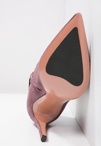 Fioletowy zamszowy but na wysokim obcasie z ostrym czubkiem, z czarną gumową podeszwą i beżowym akcentem na obcasie. Czysta, smukła sylwetka.