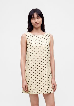 Mujer joven con cabello largo y oscuro, vestida con un vestido sin mangas de color crema con lunares marrones, de pie frente a un fondo claro y liso.