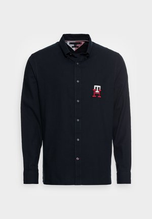 Czarna koszula z długim rękawem, kołnierzykiem i guzikami, z czerwono-białym haftowanym logo "TH" po lewej stronie klatki piersiowej.